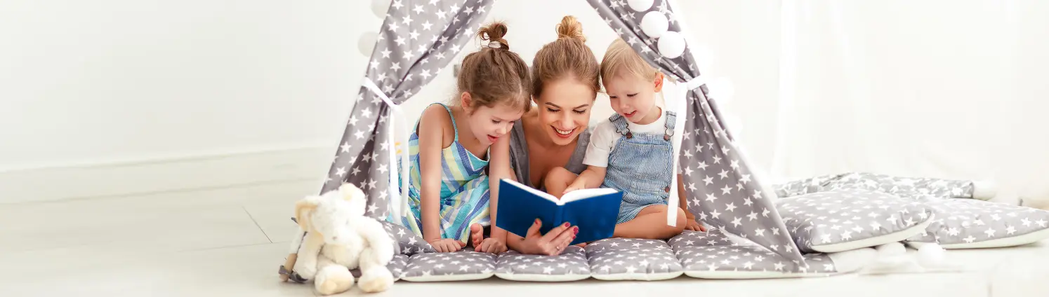 Eine Frau liegt mit zwei Kindern in einem Spielzeugzelt und liest aus einem blauen Buch vor