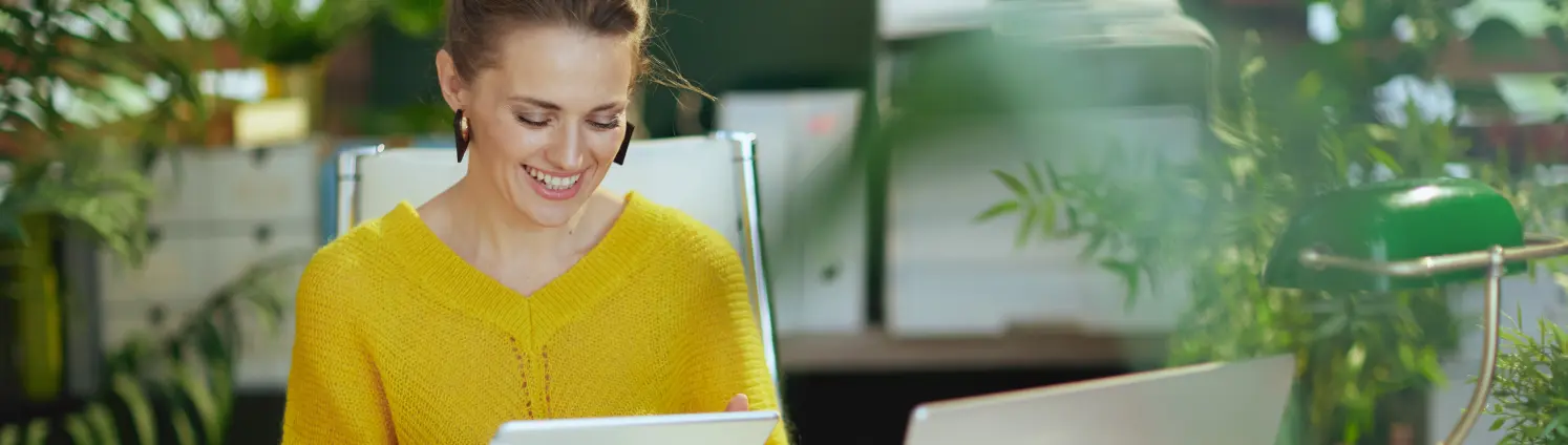 Eine Frau im gelben Pullover sitzt vor dem Laptop und hält ein Tablet in der Hand